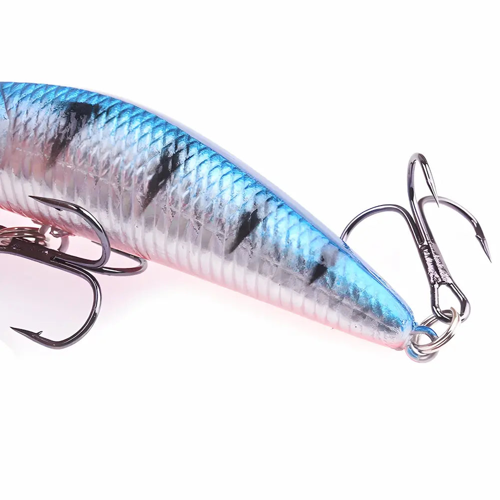 A imagem mostra um close-up de uma isca de pesca artificial. A isca tem um corpo prateado com listras pretas e detalhes em azul na parte superior. Ela possui dois anzóis triplos de cor escura. O fundo é branco, destacando a textura escamosa e as cores vibrantes da isca.